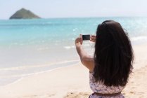 Rückansicht einer Frau, die am Strand ein Foto mit dem Handy anklickt — Stockfoto