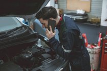 Mecánico hablando en un teléfono móvil mientras examina el coche en el garaje de reparación - foto de stock