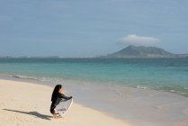 Femme accroupie avec planche de surf à la plage par une journée ensoleillée — Photo de stock
