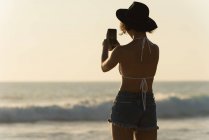 Vista trasera de la mujer haciendo clic en las fotos con teléfono móvil en la playa - foto de stock