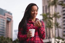 Souriant belle femme utilisant un téléphone mobile tout en prenant un café dans la rue — Photo de stock