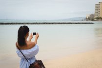 Вид ззаду жінки, що клацає фотографією моря з мобільним телефоном — стокове фото