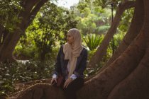 Thoughtful hijab woman sitting on tree root in garden — Stock Photo
