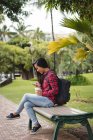Улыбающаяся женщина сидит на скамейке и пользуется мобильным телефоном в парке — стоковое фото