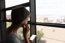 Reife Geschäftsfrau hört über Kopfhörer zu und schaut durchs Fenster ins Büro — Stockfoto
