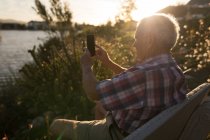 Homme âgé prenant des photos avec téléphone portable au bord de la rivière pendant le crépuscule — Photo de stock