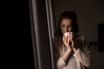 Удумлива жінка має каву, дивлячись через вікно вдома — стокове фото