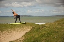 Surfeur avec planche de surf effectuant des exercices d'étirement à la plage par une journée ensoleillée — Photo de stock