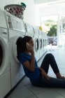 Young woman sitting on the floor and talking on the phone at laundromat — Stock Photo