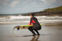 Surfer mit Surfbrett hockt an einem sonnigen Tag am Strand — Stockfoto