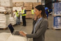 Female supervisor talking on mobile phone while using laptop in warehouse — Stock Photo