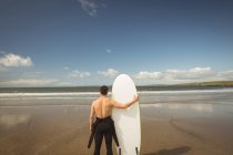 Vista posteriore del surfista con tavola da surf che guarda il mare dalla spiaggia — Foto stock