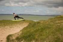 Surfista com prancha realizando exercícios de alongamento na praia em um dia ensolarado — Fotografia de Stock