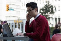 Jovem falando no telefone celular enquanto usa laptop no café ao ar livre — Fotografia de Stock