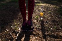 Sección baja de la atleta femenina con zapatillas de deporte de pie junto a la botella de agua en el bosque - foto de stock