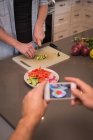 Людина фотографує жінку, готуючи салат на кухні вдома — стокове фото