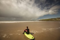 Серфер, сидящий на доске для серфинга на пляже и смотрящий на море в солнечный день — стоковое фото