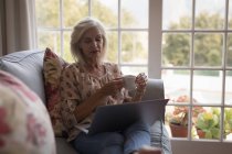 Donna anziana che prende il caffè mentre usa il computer portatile sul divano a casa — Foto stock