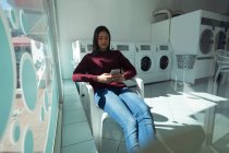Giovane donna che utilizza il suo telefono in attesa di lavanderia — Foto stock