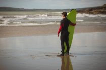 Surfista con tabla de surf mirando al mar desde la playa - foto de stock