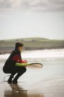 Surfeur avec planche de surf accroupi à la plage par une journée ensoleillée — Photo de stock