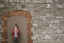 Молодий чоловік стоїть на кам'яній стіні і використовує свій мобільний телефон — стокове фото