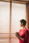 Mulher olhando através da janela enquanto toma café na sala de estar em casa — Fotografia de Stock