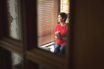 Жінка дивиться крізь вікно, маючи каву у вітальні вдома — стокове фото