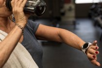 Sección media de la mujer madura revisando el tiempo mientras bebe agua en el gimnasio - foto de stock
