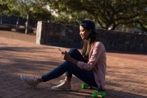 Young woman sitting on the skateboard using mobile phone — Stock Photo
