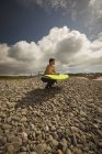 Surfista con tavola da surf accovacciata su ciottoli in spiaggia — Foto stock