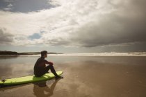 Seitenansicht eines Surfers, der am Strand auf dem Surfbrett sitzt und aufs Meer blickt — Stockfoto