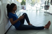 Молодая женщина сидит на полу и использует свой телефон в прачечной — стоковое фото