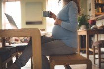 Mujer embarazada usando el ordenador portátil mientras toma café en casa - foto de stock