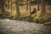 Beautiful young female hiker sitting near river coast in the forest — Stock Photo