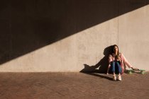 Mulher com telefone celular e skate sentado contra uma parede em um dia ensolarado — Fotografia de Stock
