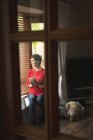 Woman looking through window while having coffee in living room at home — Stock Photo