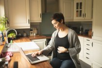 Вагітна жінка використовує ноутбук на кухні вдома — стокове фото