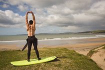 Surfer with surfboard performing stretching exercise at beach on a sunny day — Stock Photo