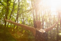 Frau mit ausgebreiteten Händen steht unter grellem Sonnenlicht im Wald — Stockfoto