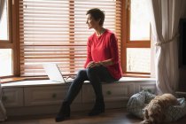 Donna che guarda attraverso la finestra durante l'utilizzo di laptop in soggiorno a casa — Foto stock