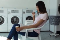Nachdenkliche Frau mit Buch und Kaffeetasse sitzt am Waschsalon — Stockfoto