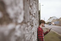 Young man using his mobile phone against a stone wall near the street — Stock Photo