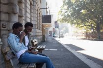Fratelli gemelli che utilizzano il telefono cellulare mentre si rilassano sulla panchina in città — Foto stock