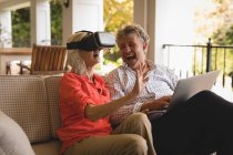 Старшая пара, использующая ноутбук и испытывающая VR гарнитуру в крыльце дома — стоковое фото