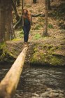 Женщина-туристка, идущая по упавшему стволу дерева через реку в лесу — стоковое фото