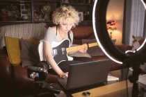 Blogueuse avec guitare utilisant un ordinateur portable dans le salon à la maison — Photo de stock
