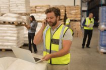 Male supervisor talking on mobile phone while using laptop in warehouse — Stock Photo