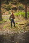 Schöne junge Wanderin, die in der Nähe der Flussufer im Wald steht — Stockfoto