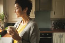 Mujer mirando hacia otro lado mientras toma café en la cocina en casa - foto de stock